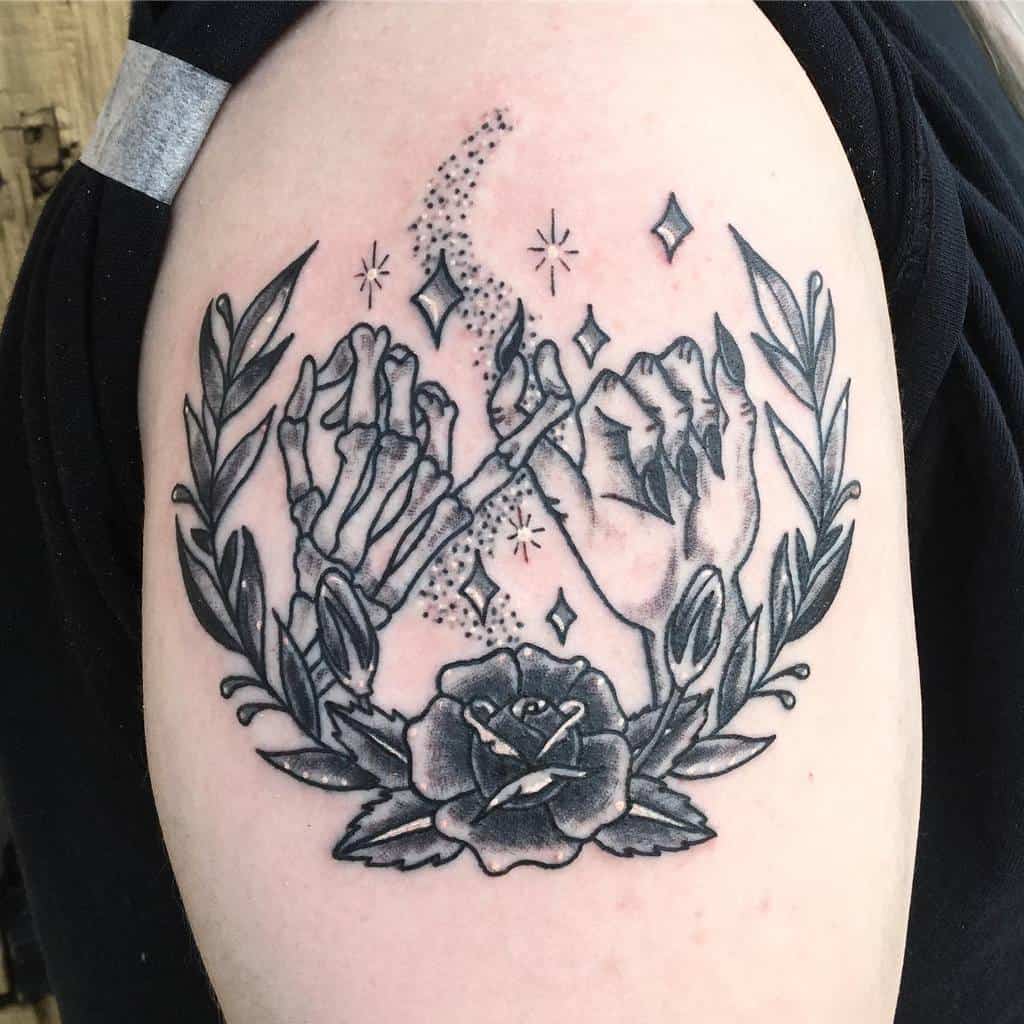 Black and grey tattoo of a pinky promise between a human and skeleton hand, surrounded by laurel leaves, a rose, and sparkling details