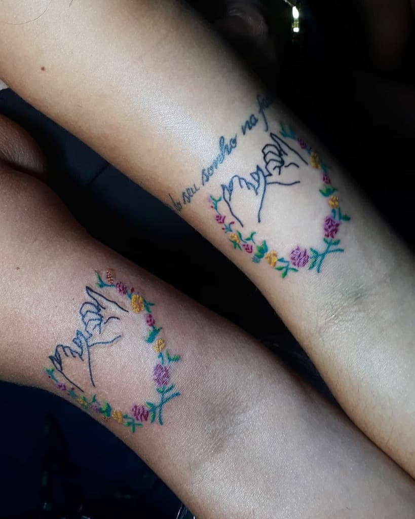 Two arms with matching tattoos of pinky promise hands inside floral wreaths, one with a cursive text above the design