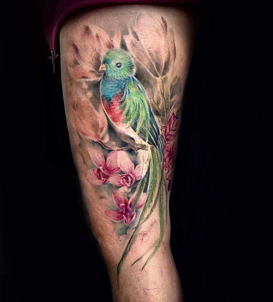 Realistic watercolor-style quetzal tattoo on the thigh, featuring a vibrant green and red bird perched among soft pink flowers with a natural, artistic background