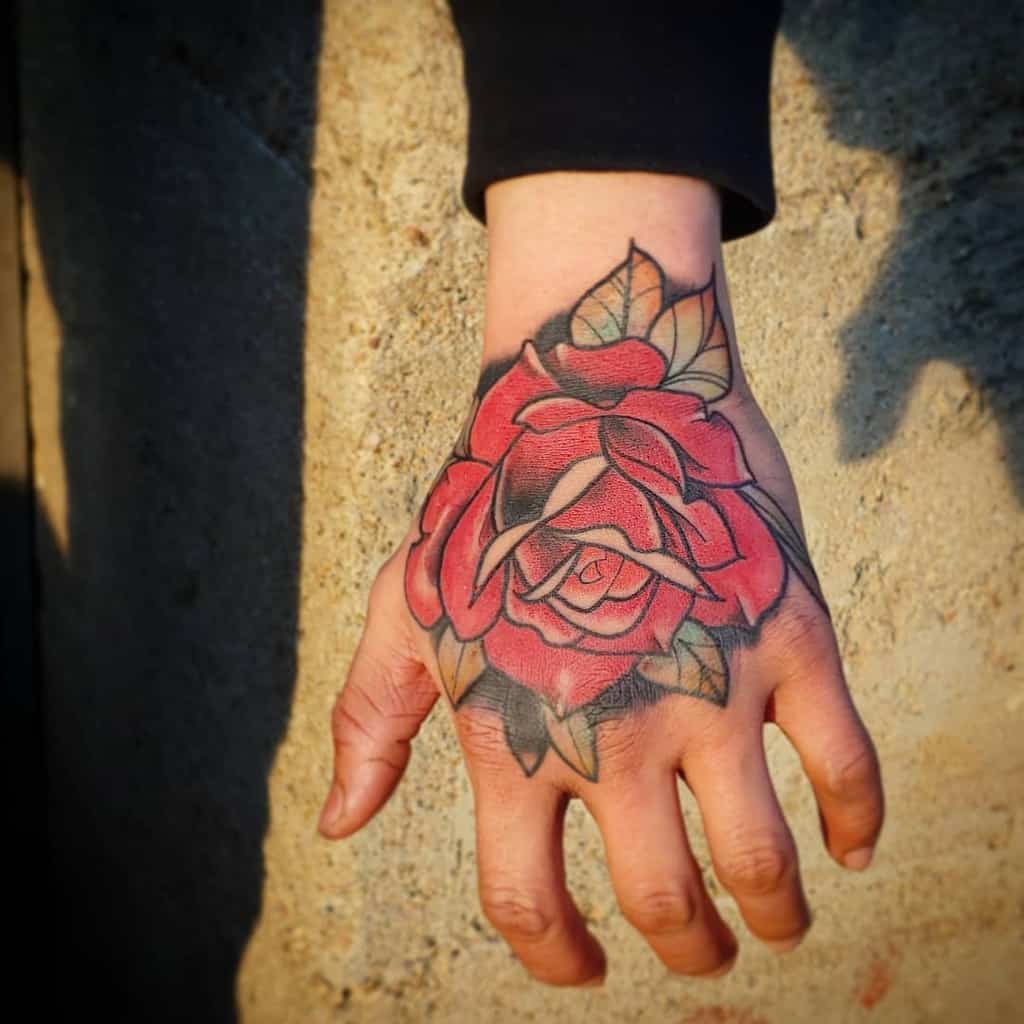 A vibrant traditional-style red rose tattoo on the hand, surrounded by bold black outlines and green leaves, capturing a classic tattoo aesthetic