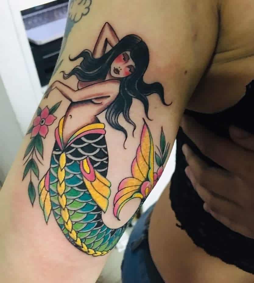 Bright and bold mermaid tattoo in traditional style, featuring a black-haired siren with vivid green and yellow scales, intricate floral accents, and a charming vintage aesthetic