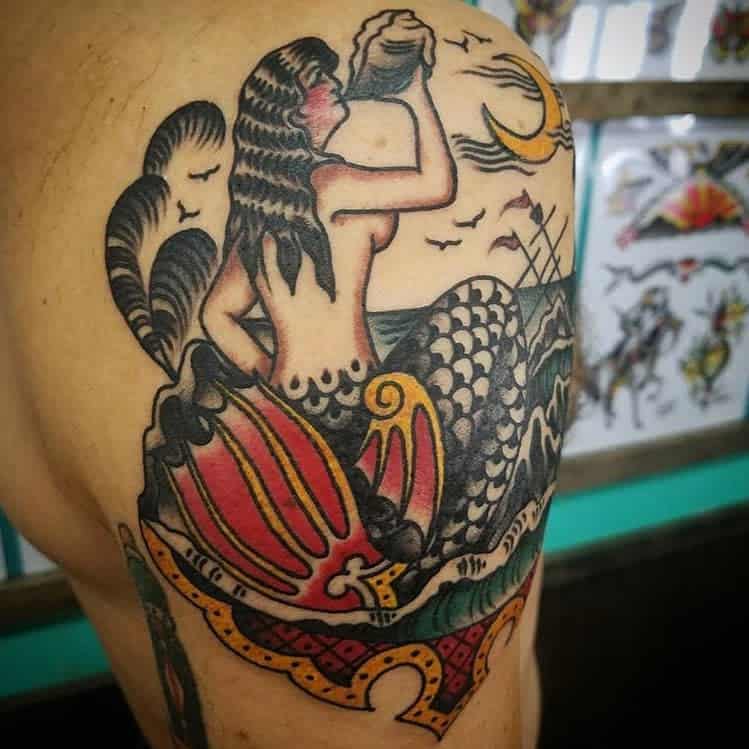 Tattoo of a vintage-style siren on a shoulder, with waves, a ship, and a shell