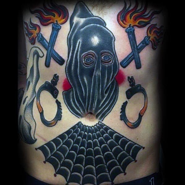 Bold executioner tattoo featuring a hooded figure with piercing eyes, crossed torches, handcuffs, and a spiderweb in neo-traditional style.