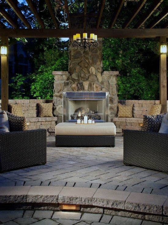 Cozy outdoor seating area with a fireplace, wicker furniture, and overhead lighting under a pergola