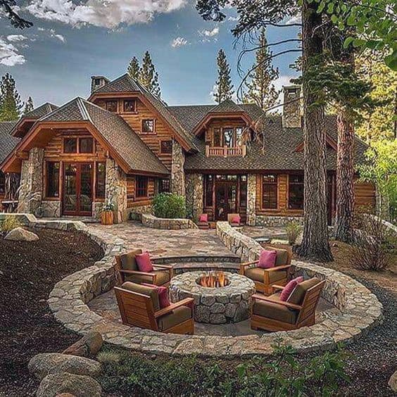 Rustic cabin surrounded by trees, featuring a stone patio with a fire pit and wooden chairs