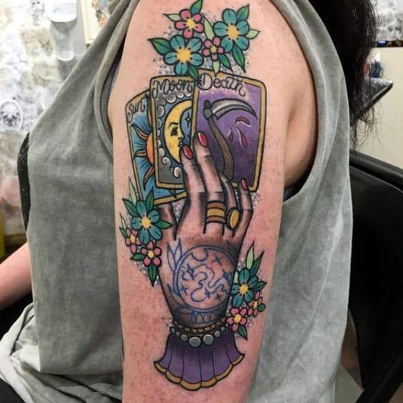 Colorful tattoo of a mystical hand holding tarot cards featuring the sun, moon, and death, surrounded by floral details on the upper arm.