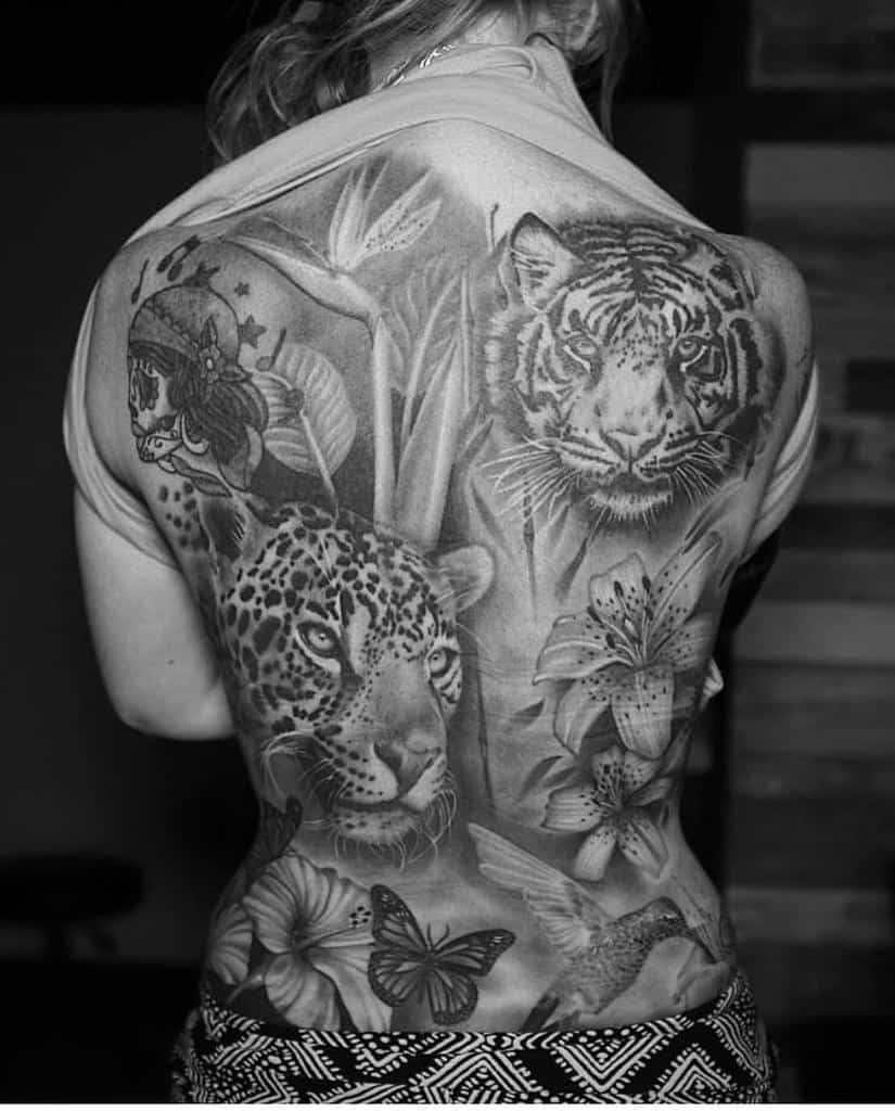 A detailed black-and-white back tattoo featuring a fierce jaguar and tiger, surrounded by lilies, butterflies, and a hummingbird, creating a vibrant, natural scene.