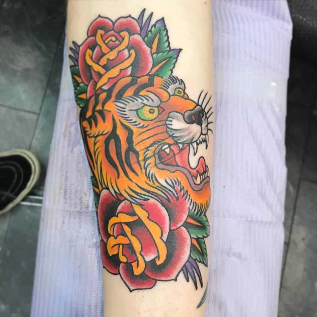 A traditional tattoo of a roaring tiger with vibrant orange and black stripes, surrounded by red roses, on the forearm