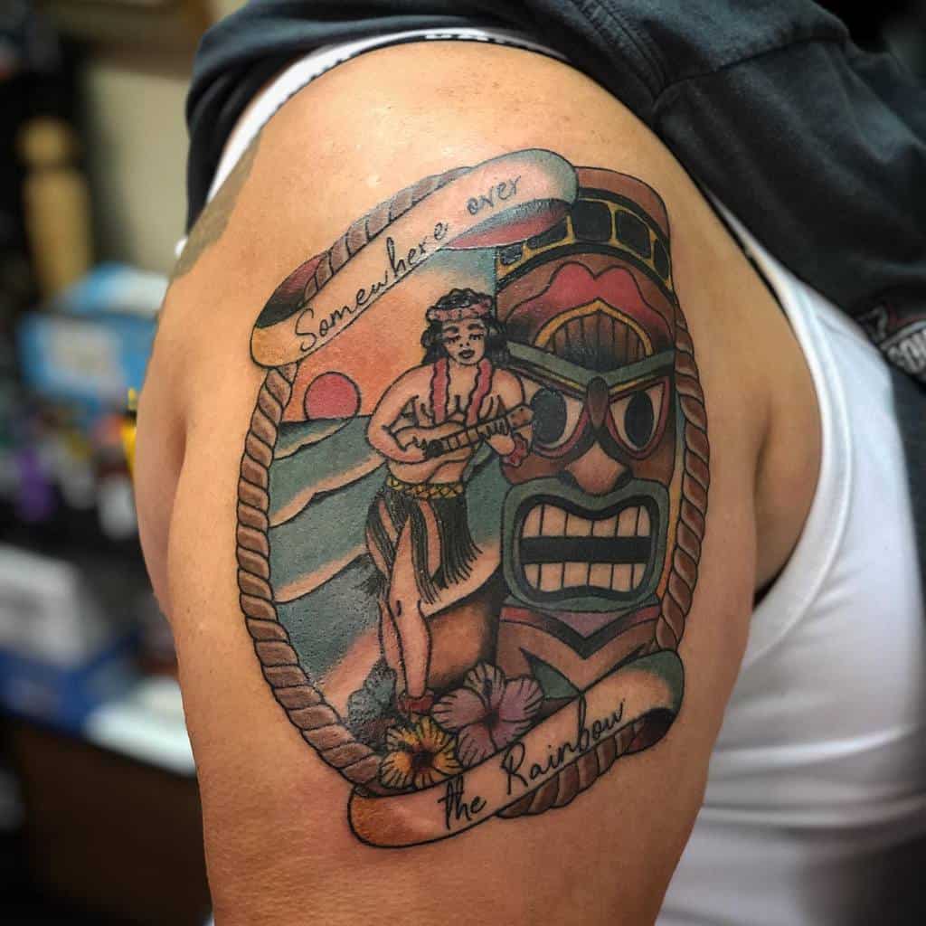 Colorful tattoo featuring a Tiki mask, a hula dancer, and a ribbon with the words 'Somewhere over the Rainbow',  flowers and blue skies, in a rope circle frame