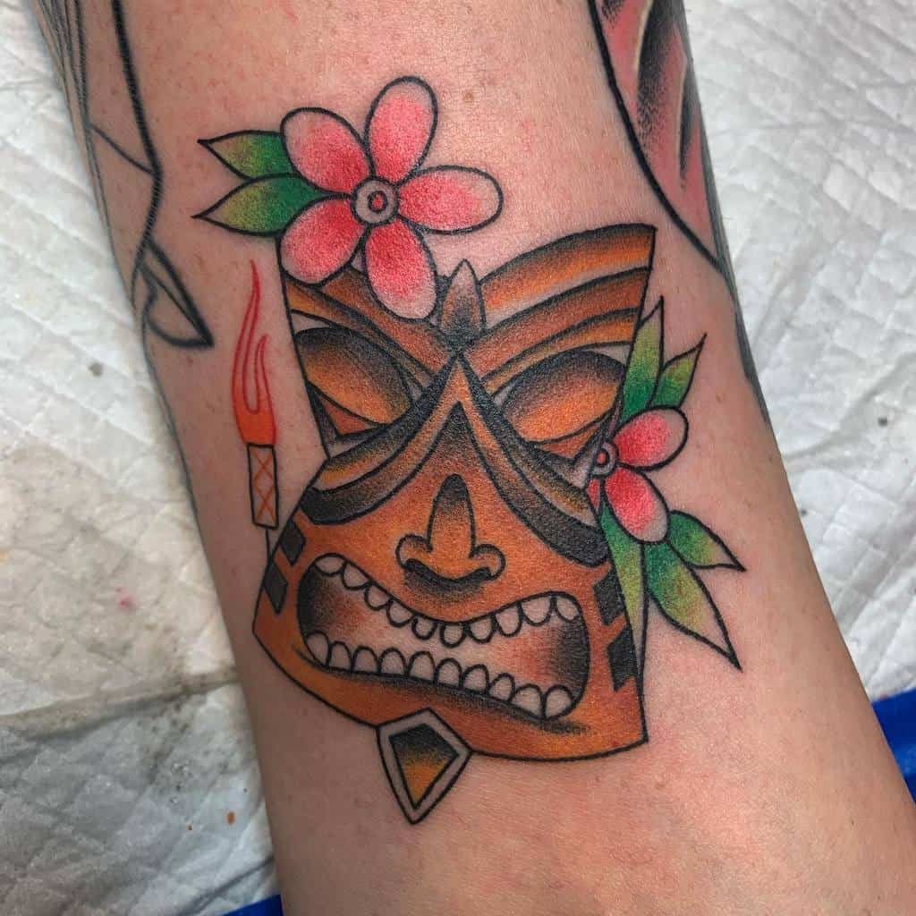 Colorful Tiki tattoo featuring a bold, angry Tiki mask with orange and black details, adorned with a pink flower, green leaves, and a lit torch