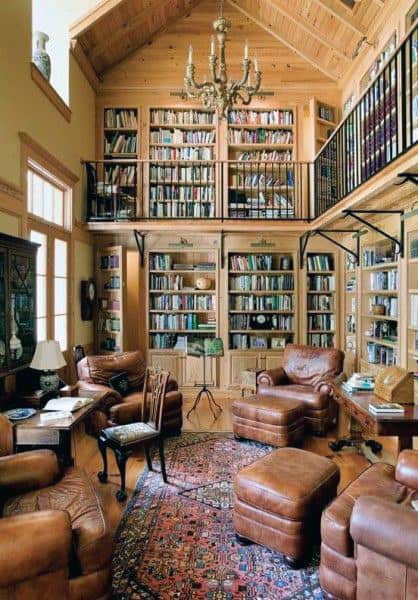 Cozy library with leather chairs, bookshelves, chandelier, and a loft, featuring warm lighting and wood accents