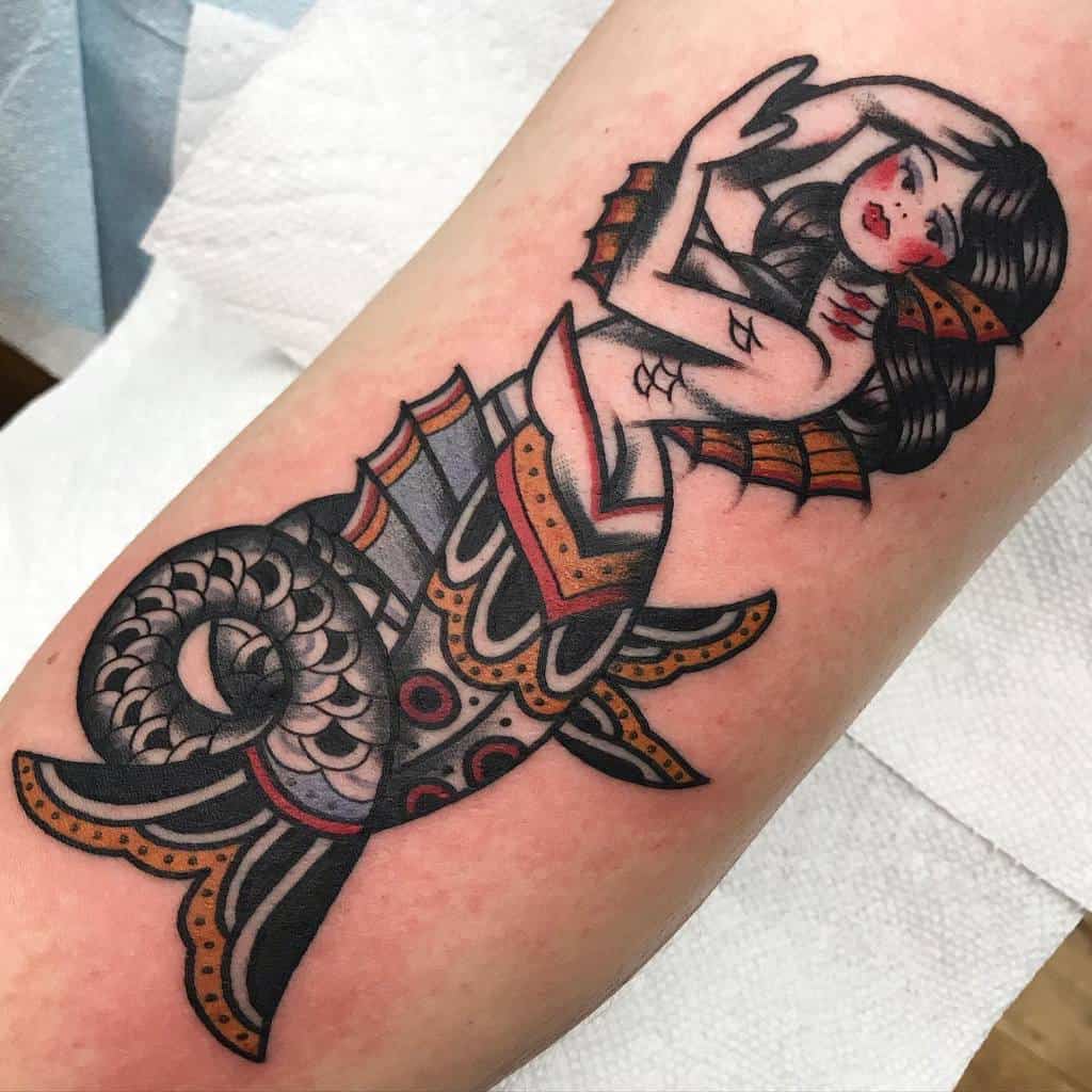 Traditional tattoo of a mermaid with a curled, ornate tail, decorative patterns, and red accents, posing with hands near her face.