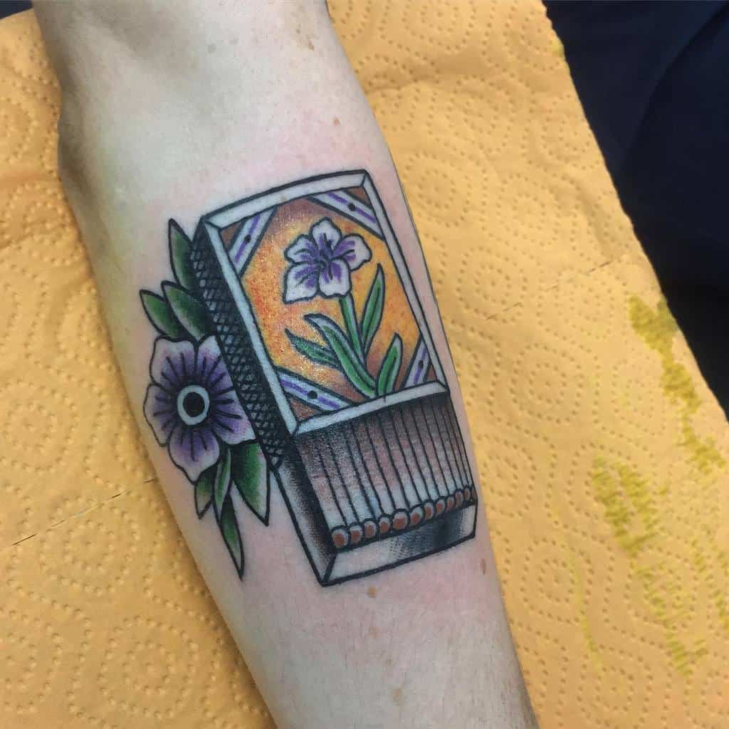 Traditional-style tattoo of a matchbox with a floral design, featuring violet flowers and green leaves, blending vintage aesthetics with bold colors