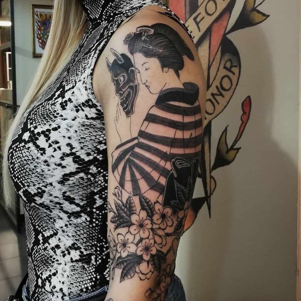 Black and gray upper arm tattoo of a Geisha holding a Hannya mask, with striped kimono details, floral elements, and intricate shading.