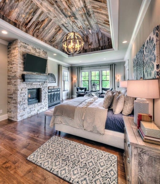 Rustic bedroom with wood-paneled ceiling, stone fireplace, and elegant natural light fixtures.
