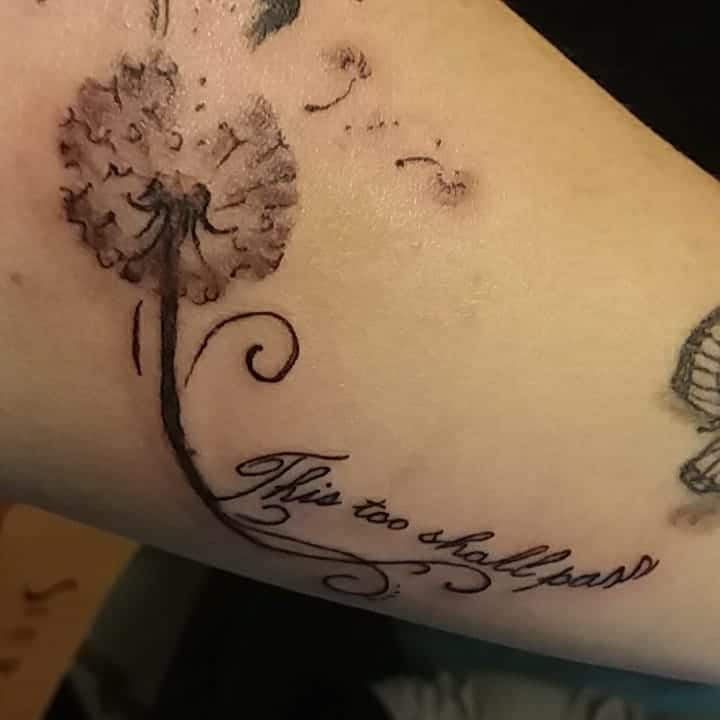 A "This too shall pass" tattoo in elegant cursive, flowing from a dandelion with seeds drifting away, symbolizing change and letting go.