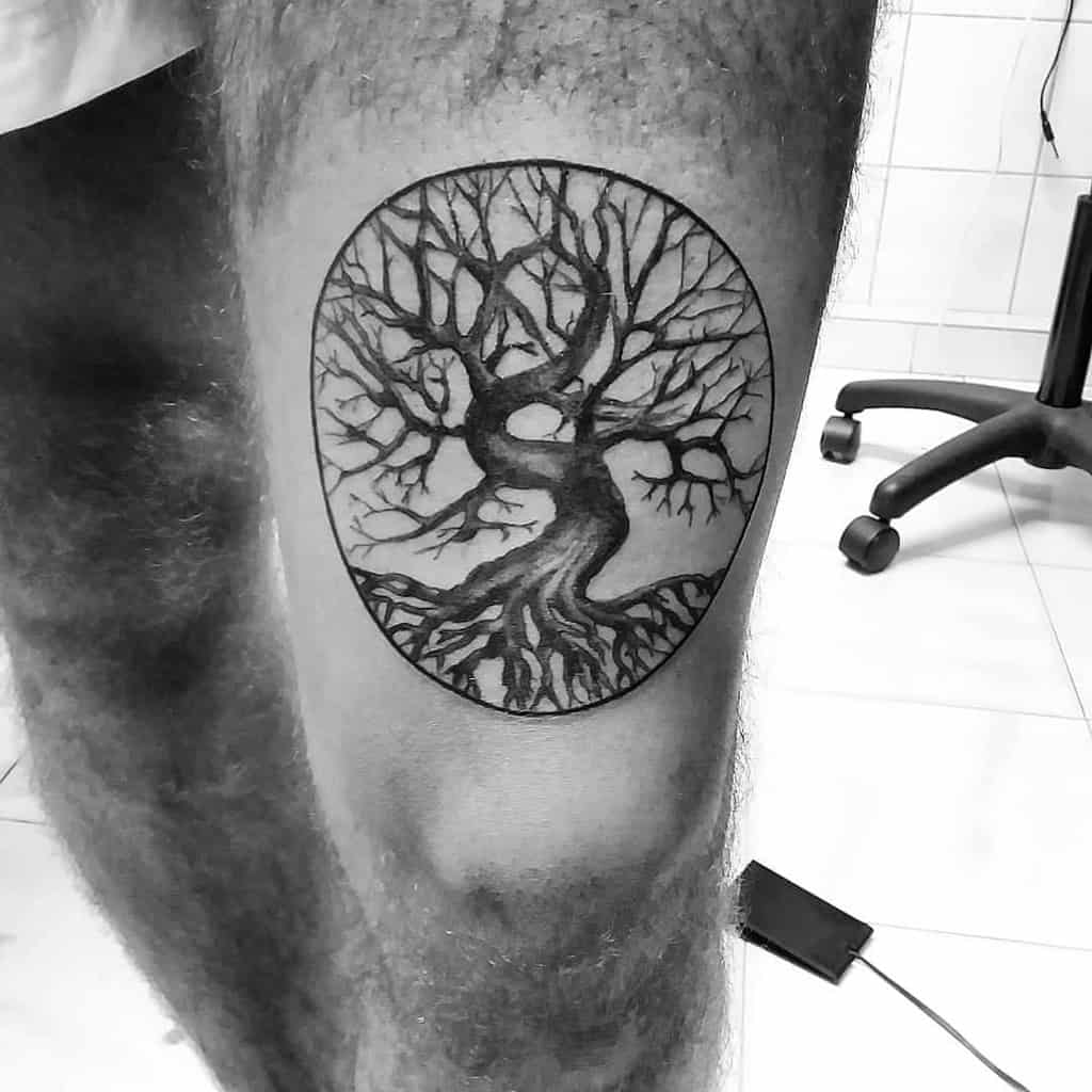 Monochrome tattoo of a tree with intricate branches and deep roots enclosed within a circular frame, representing balance and duality