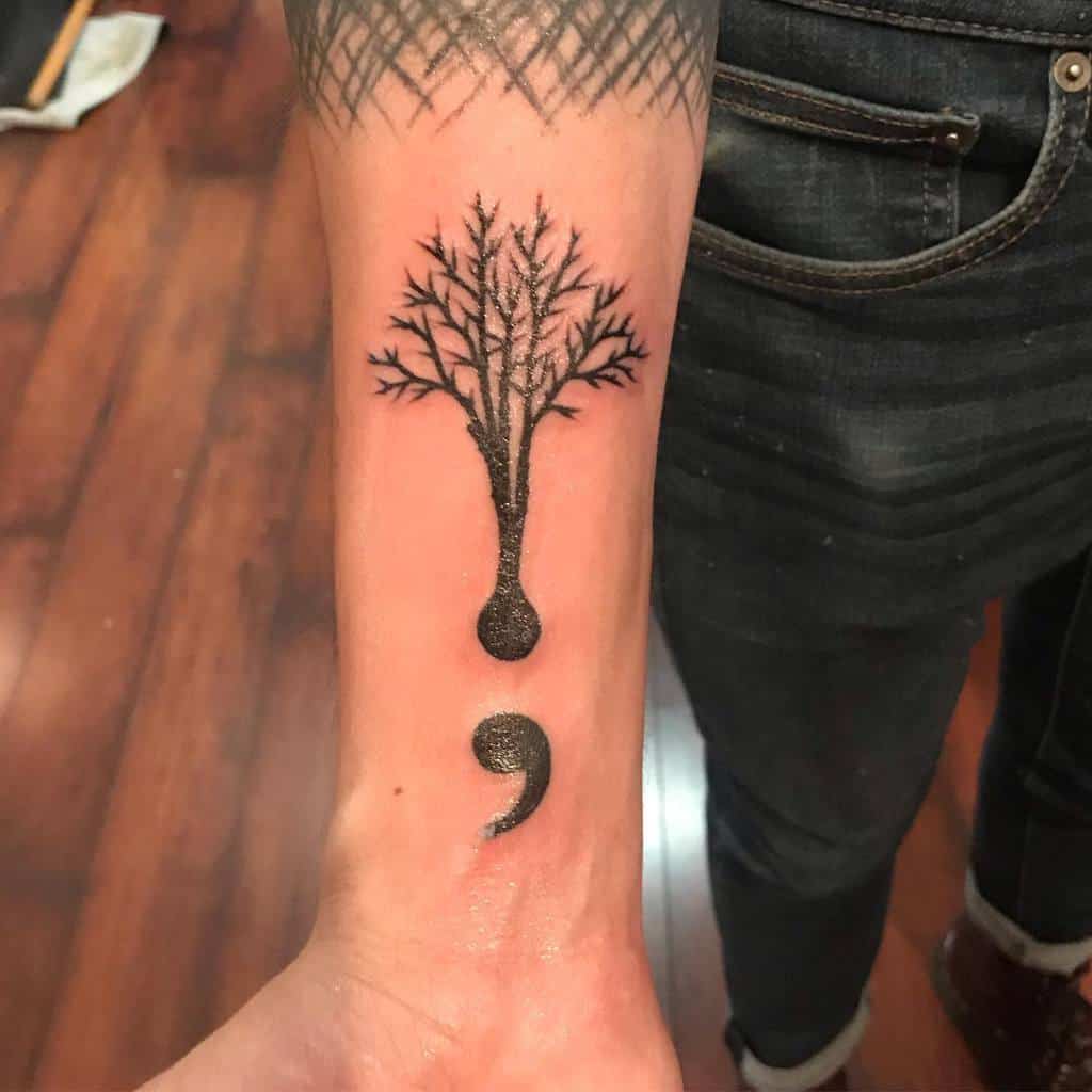 The tattoo features a tree growing from the upper part of a semicolon, symbolizing strength and resilience, with intricate details in black ink.