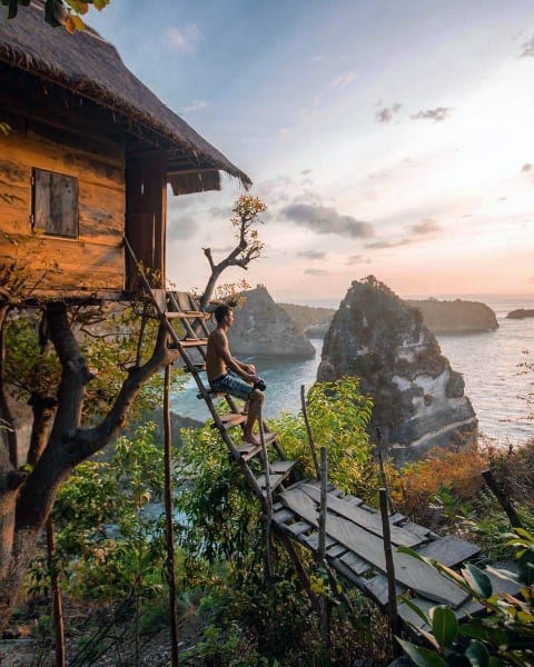 A person sits on a wooden ladder by a rustic cabin, overlooking a scenic ocean view with cliffs at sunset