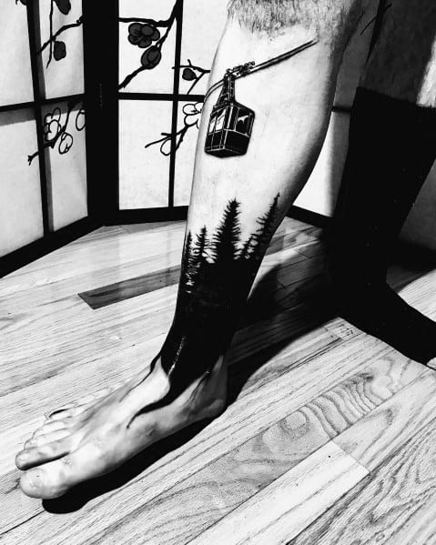 Black ink forest tattoo on the leg, featuring tall pine trees blending into the skin with a suspended cable car for a unique, nature-inspired design.
