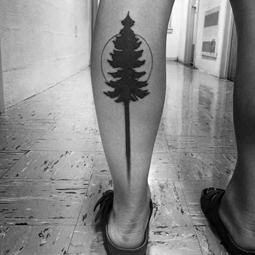Minimalist black ink pine tree tattoo on the calf, featuring a bold silhouette with a circular outline and a sharp, elongated trunk for a sleek design.