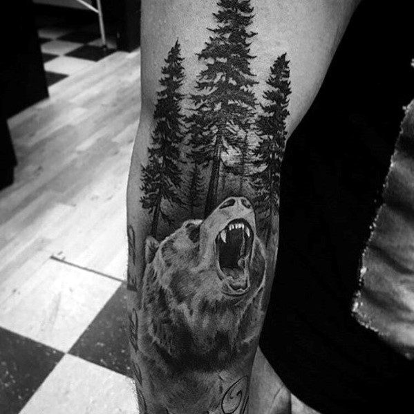 Realistic black and grey tattoo of a roaring bear with towering pine trees in the background, symbolizing strength, power, and nature.