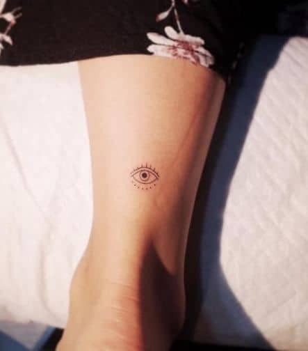 Small minimalist tattoo of an all-seeing eye with subtle radiating lines, inked in black on the ankle.
