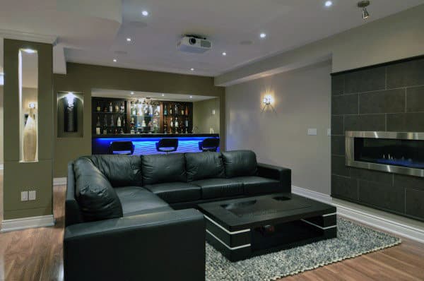 A modern basement with a black sofa, sleek bar, blue lighting, and a wall-mounted fireplace