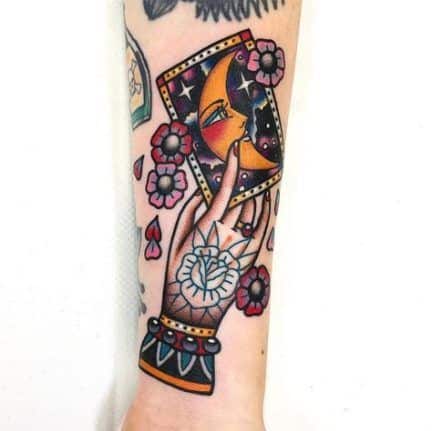 Traditional tattoo of a mystical hand holding a tarot card with a crescent moon face, surrounded by flowers and bold colors on the forearm.