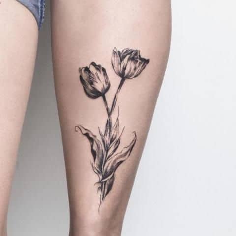 A detailed black and grey tulip tattoo on the thigh, featuring two blooming flowers with intricate shading and expressive, flowing leaves.