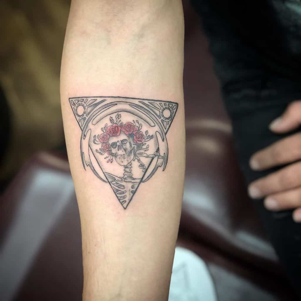 Intricate black and grey tattoo of a skull with a crown of red roses inside a geometric triangle, inked on the forearm.