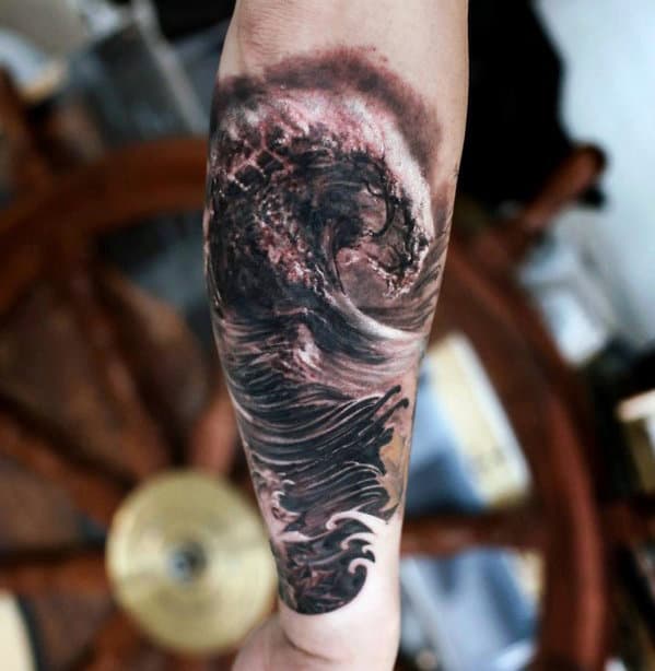 Tattoo of a wave on a forearm with a ship's wheel blurred in the background