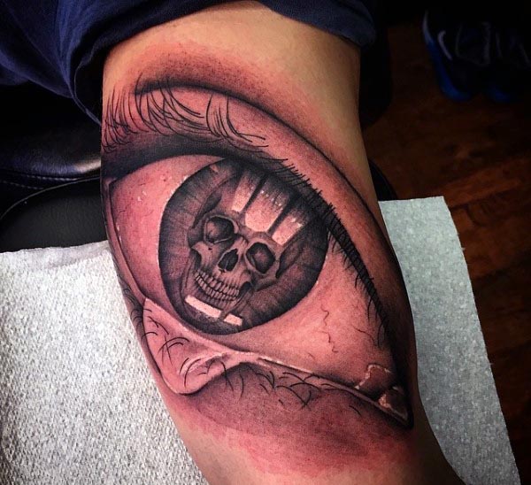 Black and grey tattoo of a realistic eye with a skull reflection in the pupil, featuring intricate shading and fine details on the inner bicep