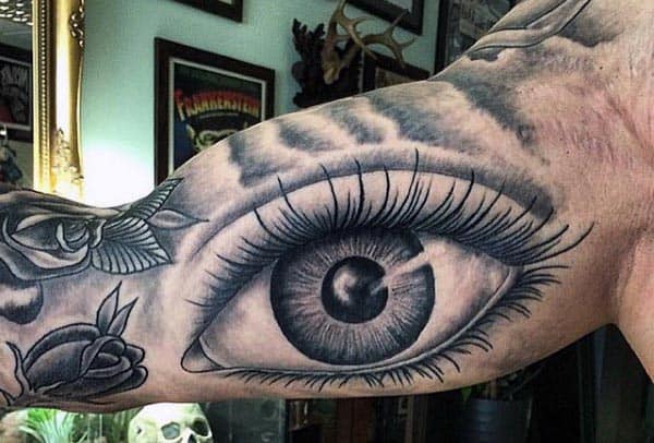 Black and grey tattoo of a highly detailed realistic eye on the inner bicep, surrounded by floral and abstract shading elements