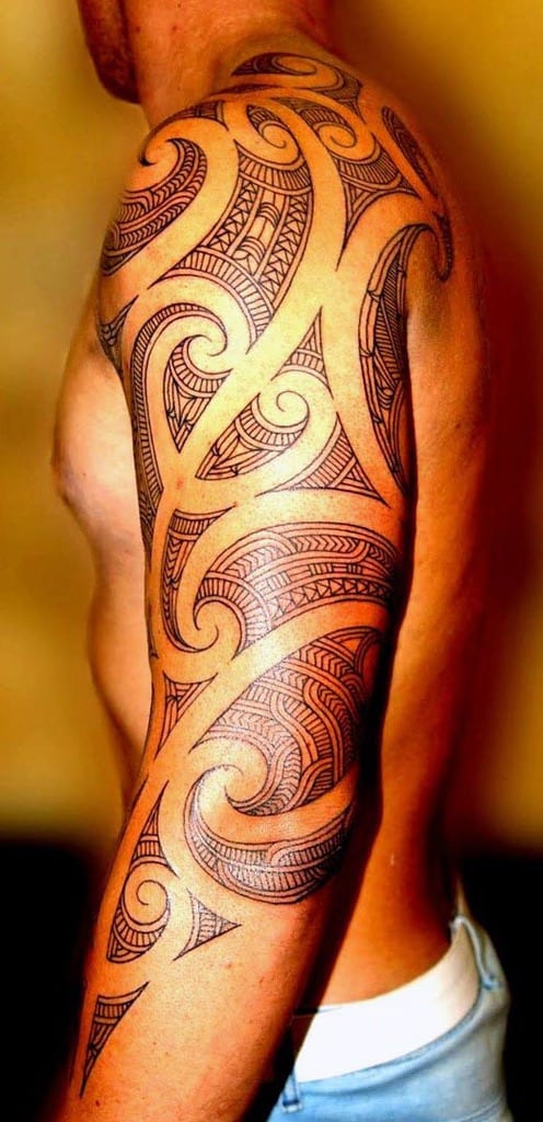 Man with intricate Polynesian tattoo on his arm and shoulder standing against a wall