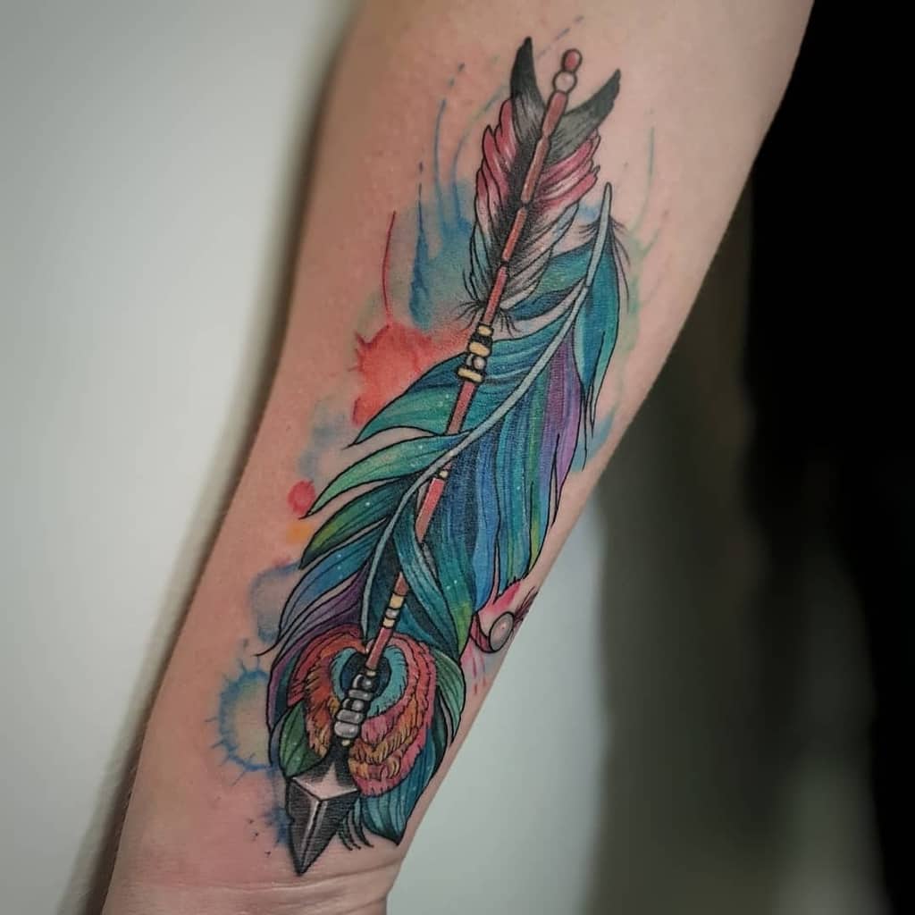 Colorful forearm tattoo of an arrow wrapped in peacock and feather elements, featuring vibrant blues, purples, and reds with watercolor splashes.