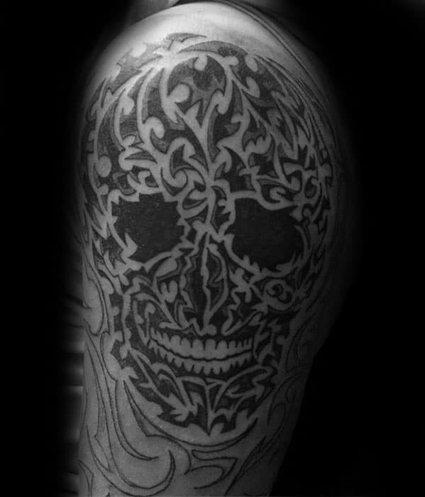 Elaborate tribal skull tattoo on upper arm with dense, interwoven patterns and dark shading, forming a bold and intense design.