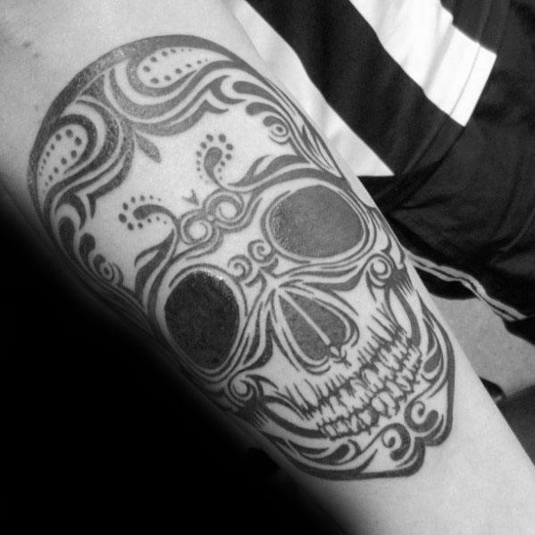 Ornate tribal skull tattoo on upper arm with swirling patterns, bold black eyes, and detailed linework for a decorative sugar skull look.