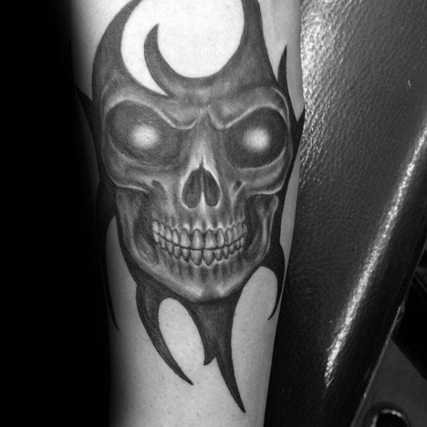 Dark tribal skull tattoo on forearm with glowing eyes, sharp flame-like edges, and intense shading for a fierce, menacing look.