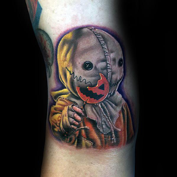 Realistic color tattoo of Sam from *Trick 'r Treat*, featuring his stitched burlap mask, orange outfit, and signature broken pumpkin lollipop for a spooky effect.