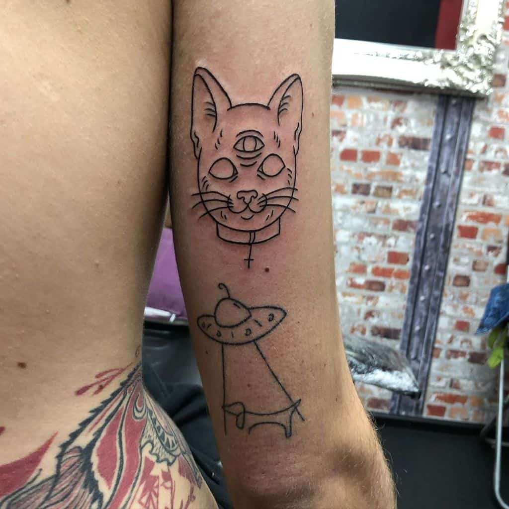 Minimalist tattoo of a three-eyed cat with a UFO beaming up a cow, inked in fine black lines on the upper arm.
