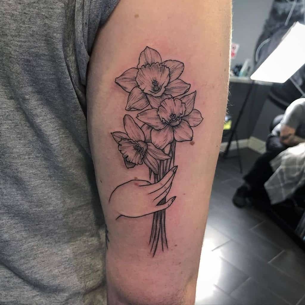 This tattoo features a beautiful bouquet of daffodils held delicately by a hand, creating a sense of elegance and grace. The fine linework in the flowers adds intricacy, while the hand complements the composition, giving the design a personal touch. The shading is subtle but adds depth to the flowers, making them pop. This design combines nature and human elements to create a unique and meaningful piece.