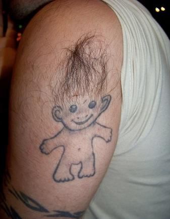 A black ink tattoo of a smiling troll doll on the upper arm, creatively using natural body hair to resemble the doll's fuzzy hair.