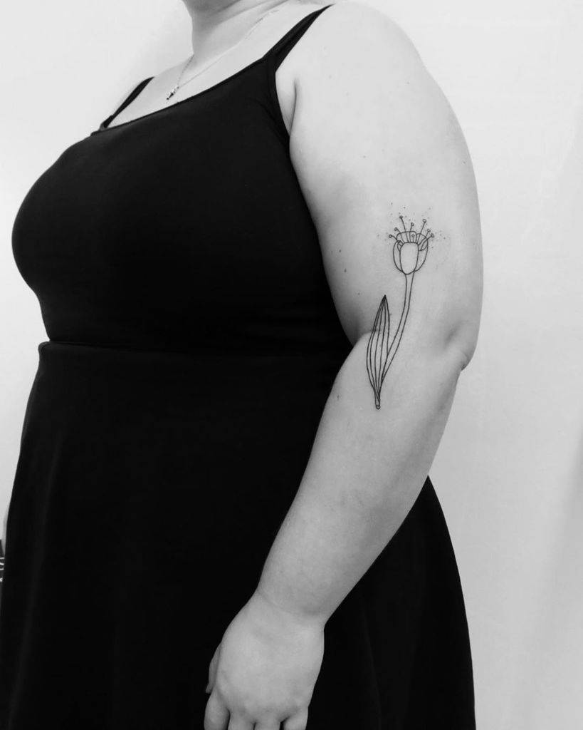 A black-line tulip tattoo on the upper arm, featuring a delicate flower with small dot accents and a slender stem and leaves.