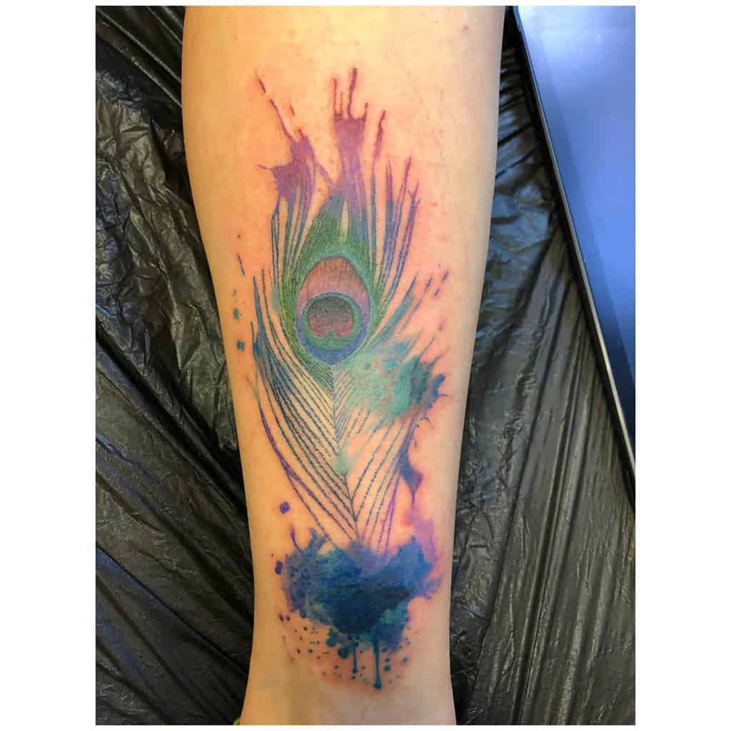 Watercolor-style peacock feather tattoo on the forearm, featuring vibrant green, blue, and purple hues with abstract ink splashes.