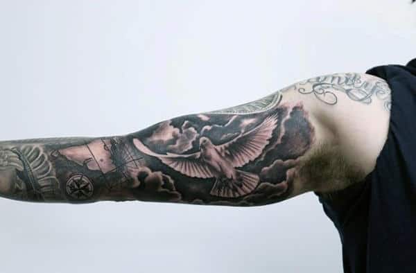 Tattooed arm featuring a dove flying through clouds, compass design, and intricate imagery