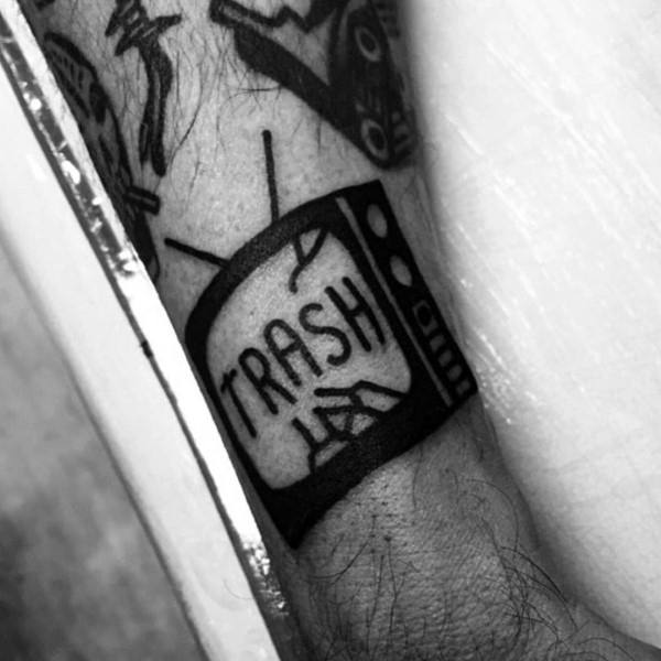 Black ink tattoo of a vintage TV with the word "TRASH" on the screen, accompanied by a hand-drawn symbol, expressing criticism of media content.