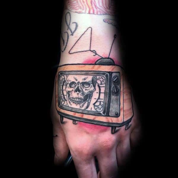 Black and grey tattoo of a vintage TV on the hand, displaying a grinning skull on the screen, with fine line details and a slightly distorted effect.