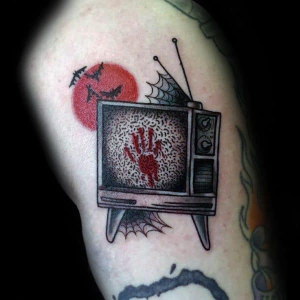 Tattoo of a vintage TV with a static screen featuring a bloody handprint, surrounded by cobwebs, a red moon, and flying bats, creating a horror theme.