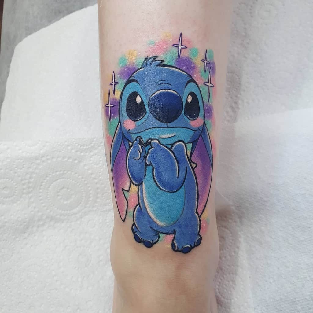 Colorful tattoo of Stitch from Lilo & Stitch with a cute expression, surrounded by pastel-colored stars and sparkles, on the forearm.